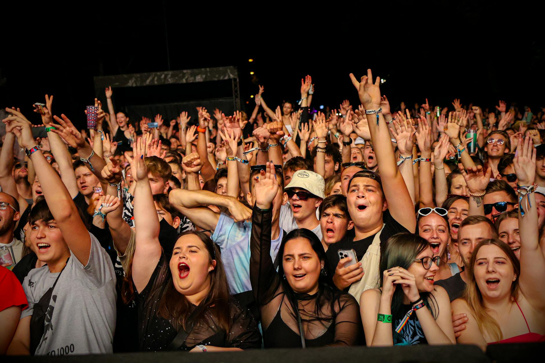 Large concert crowd with raised hands and phone lights in dark venue
