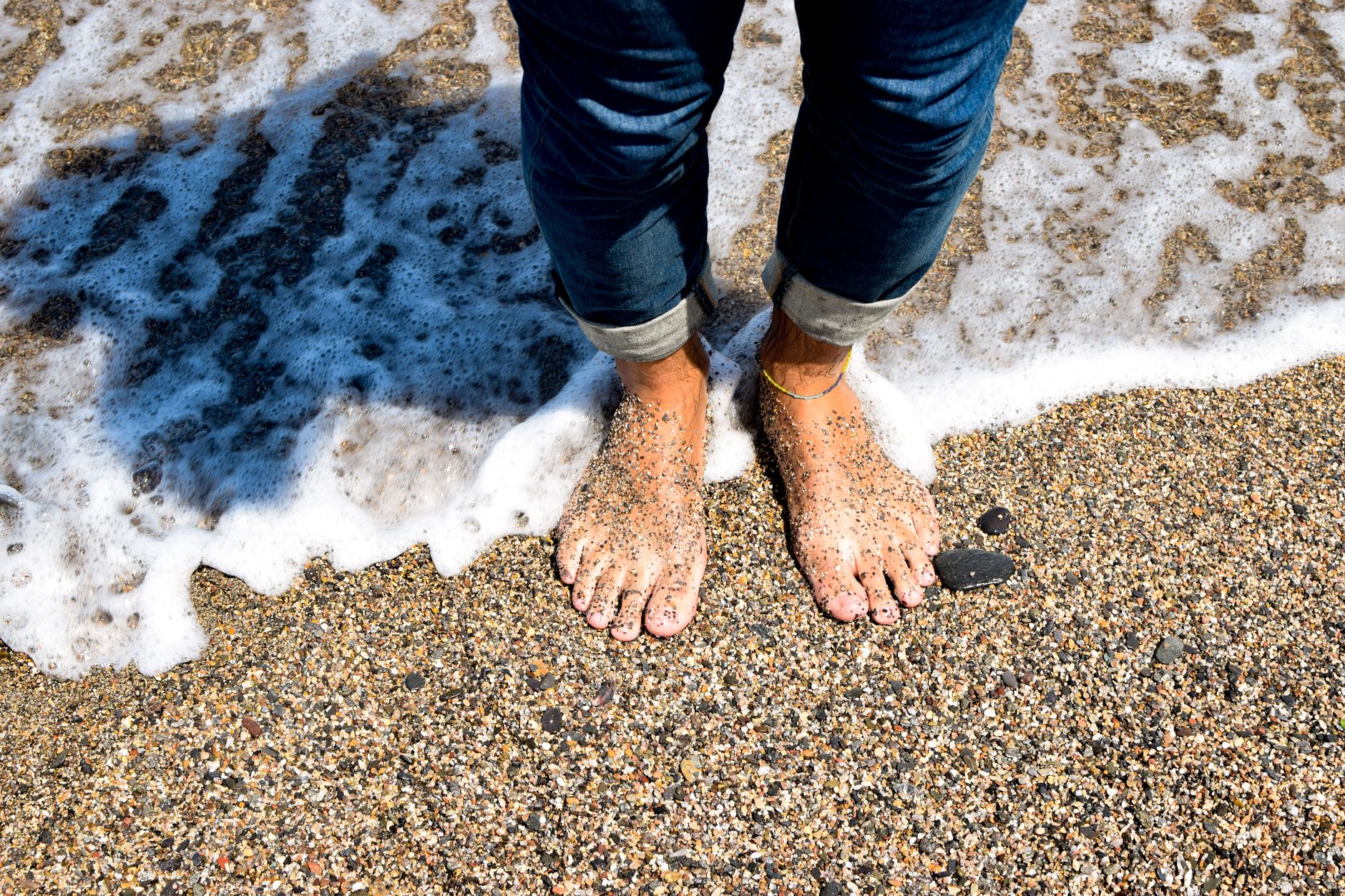 Bare feet in sand wearing layered anklets with bright gemstone charms